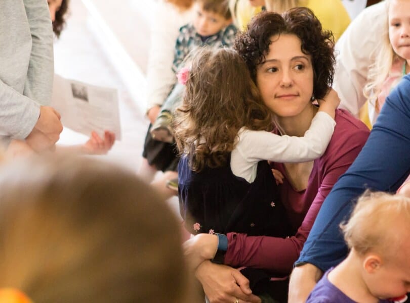A mother hugs her daughter during the children's sermon. Other parents and children surround them.