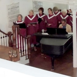 Choir members surround a piano and sing