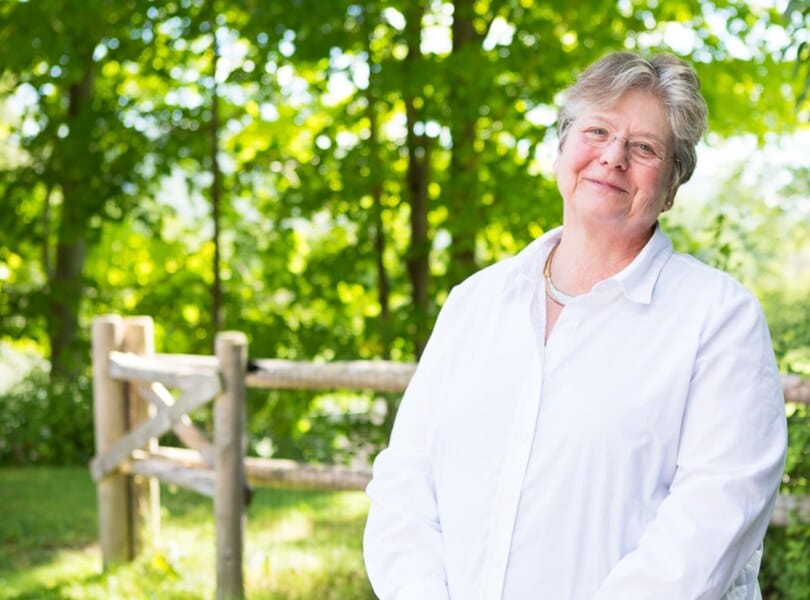 A woman clasps her hands and smiles with green trees in the background