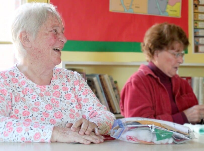 A woman crochets while another woman smiles