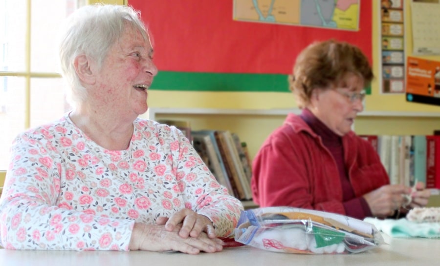 A woman crochets while another woman smiles